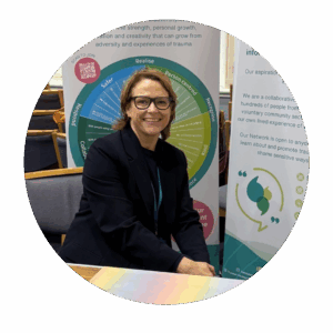 A smiling woman wearing glasses and a dark blazer sits at a table in front of two informational display banners. Behind her, one banner features a colorful circular diagram with sections labeled about trauma-informed and person-centred approaches. The setting appears to be an indoor event or conference space with chairs in the background.